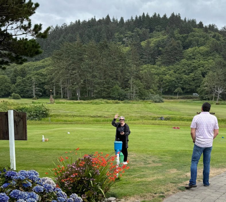 Nikki waving from a golf course practice green while Gunny looks on, with forested hills behind