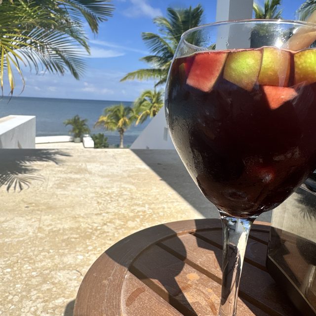 A glass of fruit-filled sangria on a poolside table, palms and the sea behind